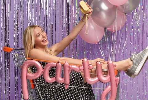 Bruden i festkjole sitter i handlevogn med ballonger og tar selfie foran glittervegg – klar for et utdrikningslag full av latter og påfunn.