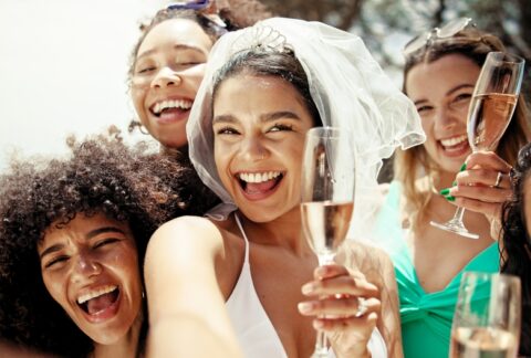 Brud og vennegjeng smiler og skåler med champagne under utdrikningslag i solen, klare for en fest full av latter og minner.