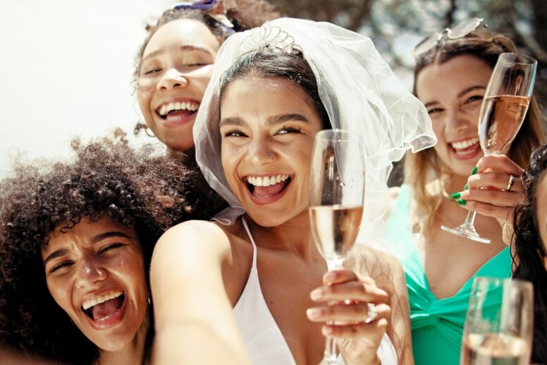 Brud og vennegjeng smiler og skåler med champagne under utdrikningslag i solen, klare for en fest full av latter og minner.